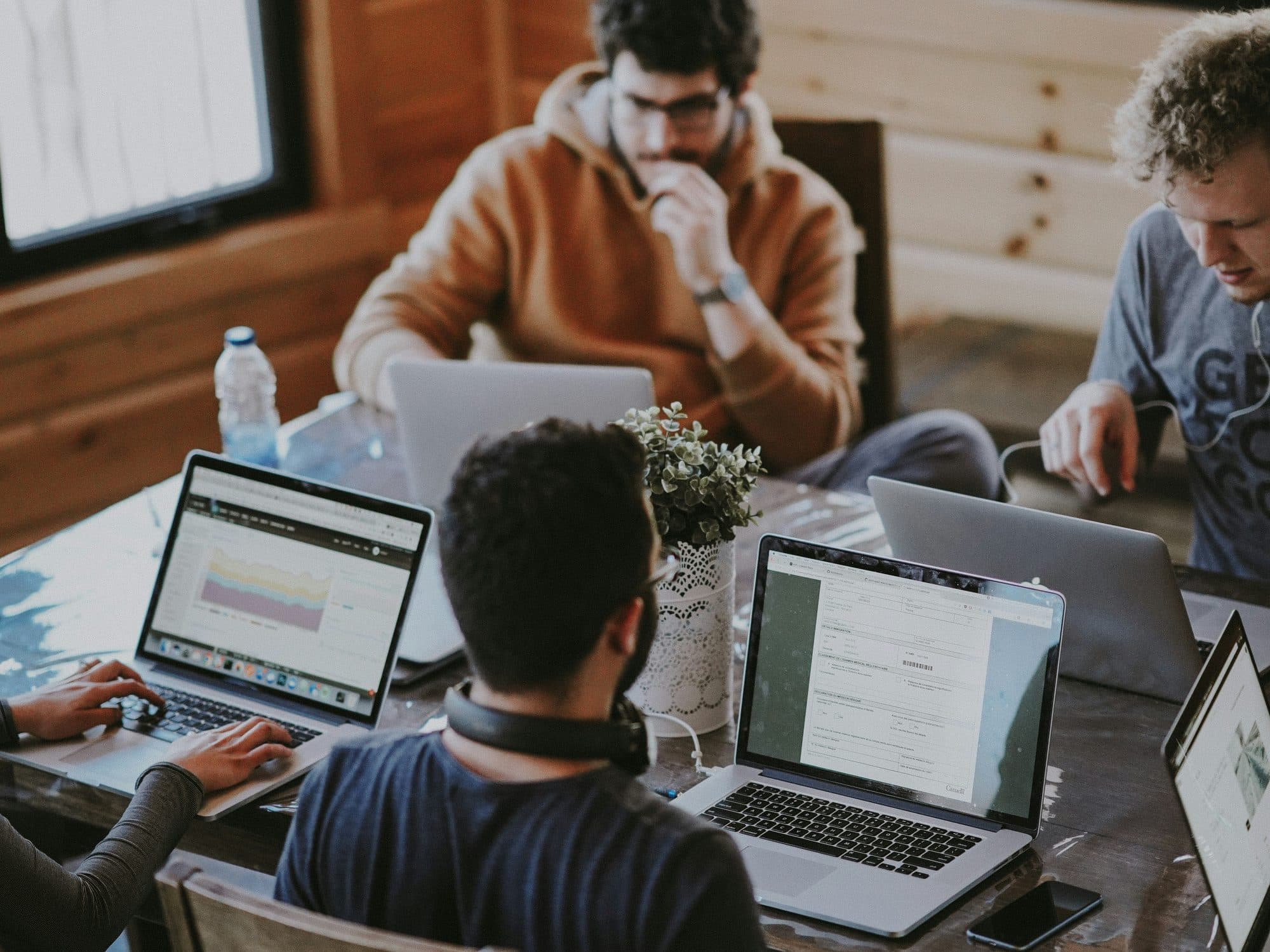 Vier Menschen arbeiten an Laptops um einen Holztisch, einer tippt, einer denkt mit der Hand am Kinn nach, auf den Bildschirmen ein Diagramm und ein Formular.